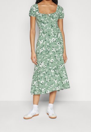 Kvinde iført en grøn midi-kjole med hvidt blomsterprint, puffede ærmer og firkantet udskæring, kombineret med hvide sneakers og sokker.