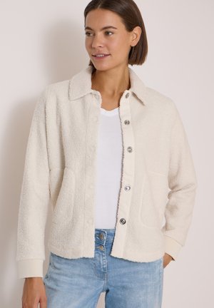 Femme portant une veste polaire crème boutonnée sur un t-shirt blanc et un jean bleu clair, debout avec une main dans la poche et regardant vers la droite.