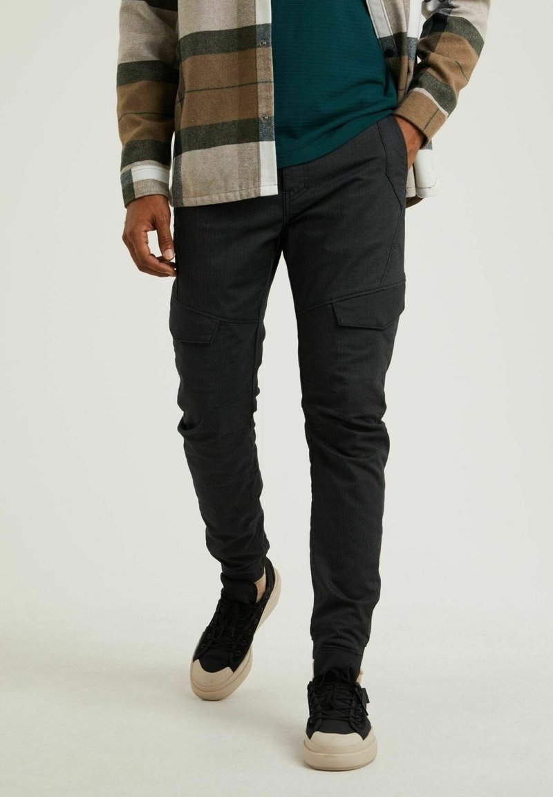 Pantalon cargo noir avec multiples poches, coupe fuselée, tissu texturé, associé à des baskets noires et beiges. Haut décontracté non visible.