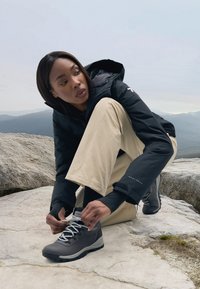 Black insulated jacket, beige pants, and gray hiking boots with white laces. Sitting on a rock, adjusting shoelaces outdoors.