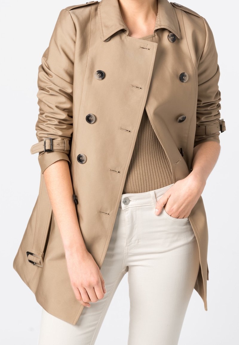 Personne portant un trench-coat beige à double boutonnage avec les manches retroussées sur un haut beige à côtes et un pantalon blanc ajusté, la main dans la poche.