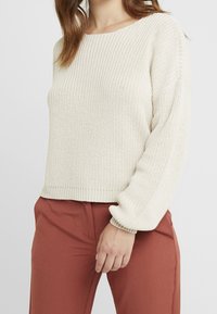 Frau trägt einen cremigen, grob gestrickten Pullover mit Ballonärmeln und rostfarbene, maßgeschneiderte Hosen, steht vor einem weißen Hintergrund.