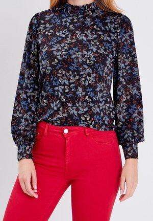 Femme portant un chemisier noir à manches longues à fleurs bleues et blanches, associé à un pantalon taille haute rouge vif.