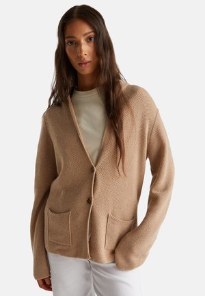 Femme portant un cardigan boutonné texturé beige avec deux poches avant sur un haut clair et un pantalon blanc, se tenant devant un fond blanc.