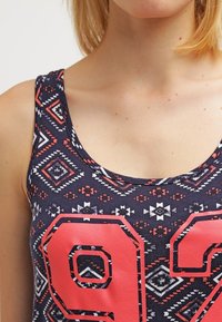 Beżowa bezrękawnikowa bluzka w kolorze granatowym z geometrycznymi wzorami w różowym i białym, z dużą czerwoną liczbą "92" na przodzie. Wykonana z miękkiej bawełny.