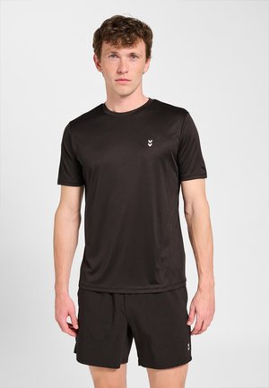 Mężczyzna ubrany w czarną sportową koszulkę z krótkim rękawem i pasujące krótkie spodenki, stojący na tle jednolitego jasnoszarego tła.