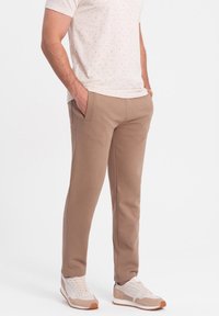 Beige Jogginghose mit glatter Textur, seitlichen Taschen und taillierter Silhouette. Getragen mit hellen Sport Sneakers.