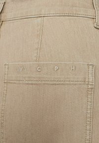Beige denim broek met een achterzak met gestikte letters "IVY CPH." De stof heeft een gladde textuur en minimale zichtbare stiksels.