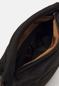 Sac en tissu noir avec une surface lisse, fermeture éclair et doublure beige ; présente un logo imprimé sur le côté intérieur.
