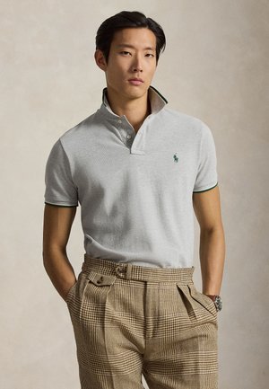 Mężczyzna o krótkich ciemnych włosach, ubrany w jasnoszarą koszulkę polo z zielonym obszyciem i beżowe spodnie w kratę z wysokim stanem, ręce w kieszeniach, neutralne tło.