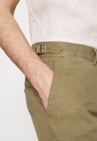 Kalhoty z olivově zeleného lnu mají boční kapsu, poutko na opasek se stříbrnou sponou a měkkou texturu, prezentované na neutrálním pozadí.