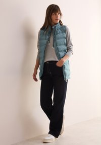 Türkisfarbene Steppweste mit Reißverschluss, graues Langarmshirt darunter, dunkle Jeans und weiße Sneakers. Glatte Textur und lässige Passform.