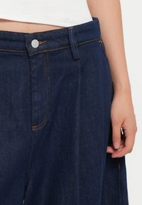 Sötétkék denim nadrág narancssárga varrással, gombos zárással és oldalsó zsebekkel rendelkezik. Az anyag sima tapintású és laza illeszkedésű.