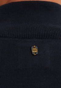 Tamno plavi pleteni pulover s rebrastim ovratnikom. Istaknuta zlatna i crna oznaka s logotipom "PME LEGEND" ušivena je na njemu.