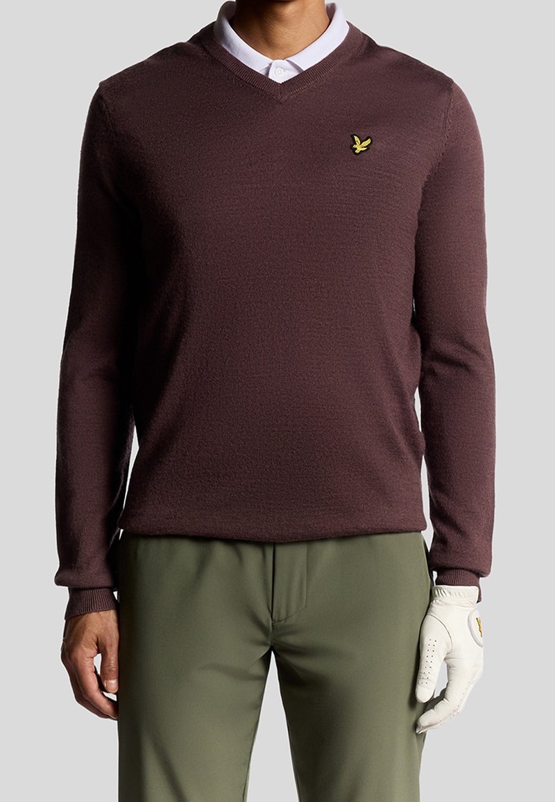 Homme portant un pull marron à col en V, une chemise blanche à col en dessous, un pantalon vert olive et un gant de golf blanc à la main droite.