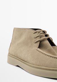 Bottines chukka en suedine beige avec un bout rond, un laçage à l'avant et une semelle lisse et plate. Présente une couture propre et des accents de design minimalistes.