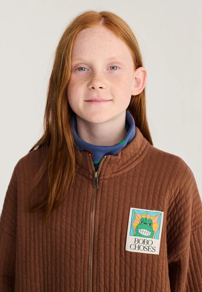 Enfant roux avec des taches de rousseur porte une veste matelassée marron avec un patch grenouille coloré portant l'inscription "BOBO CHOSES" sur la poitrine.