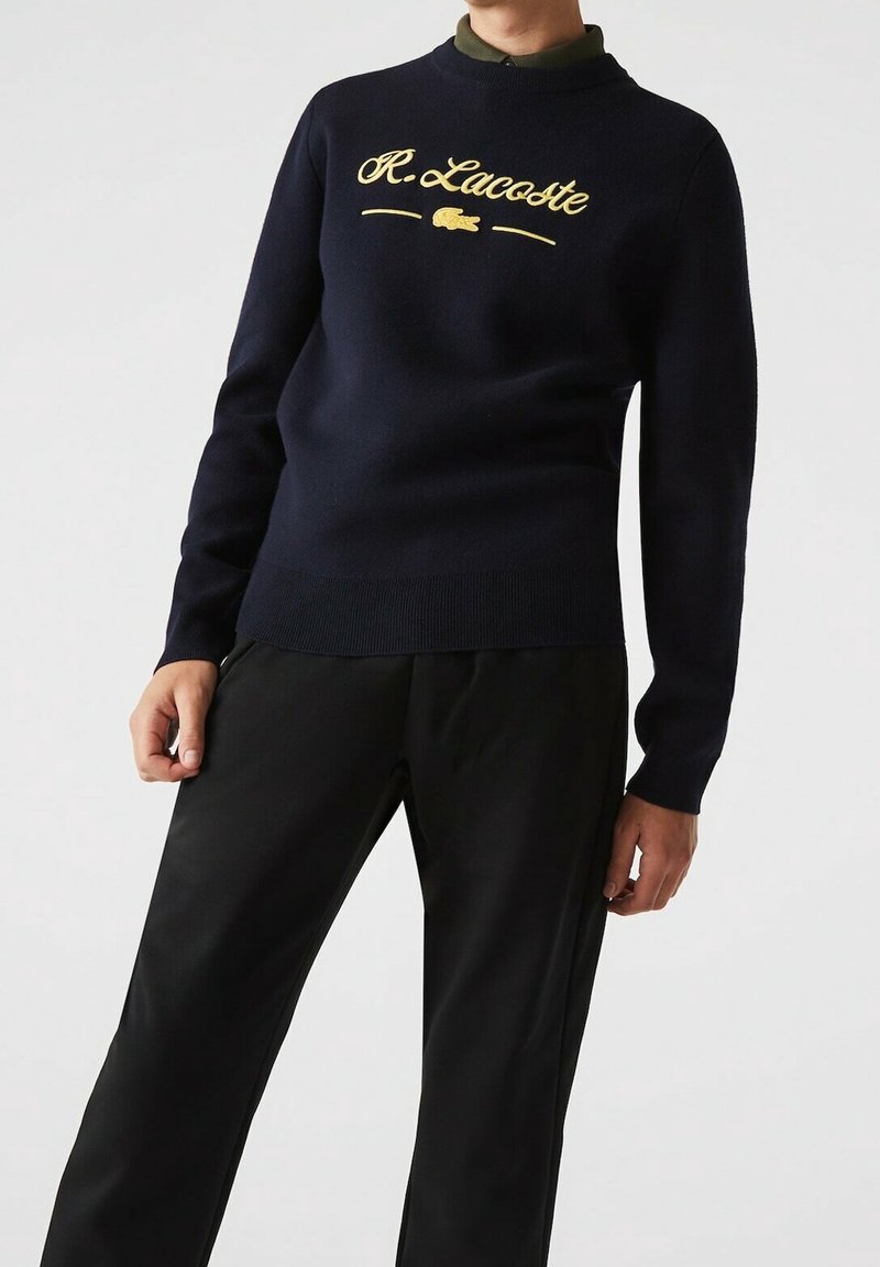Personne portant un pull noir avec le texte jaune "R. Lacoste" et le logo du crocodile, assorti à un pantalon noir sur un fond clair uni.