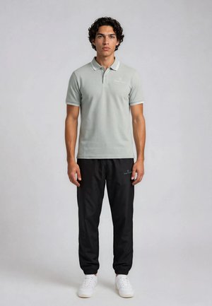 Młody mężczyzna o kręconych włosach stoi ubrany w jasnoszarą koszulkę polo, czarne spodnie dresowe i białe trampki na jednolitym szarym tle.