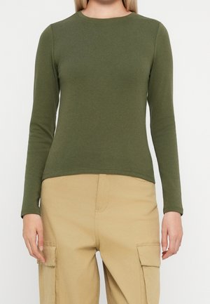 Person iført en olivengrøn langærmet skjorte og beige cargobukser med lommer, stående mod en ensfarvet lys baggrund.
