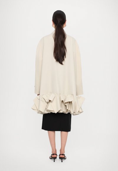 Light beige oversized jacket with ruffled hem, featuring a smooth texture. Paired with a black skirt and black heeled sandals.