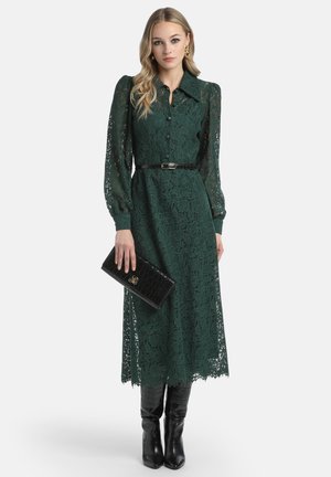 Groene kanten jurk met een kraaglijn, lange puffmouwen en een ceintuur op de taille. Gepaard met een zwarte clutch met krokodillenprint en kniehoge laarzen.