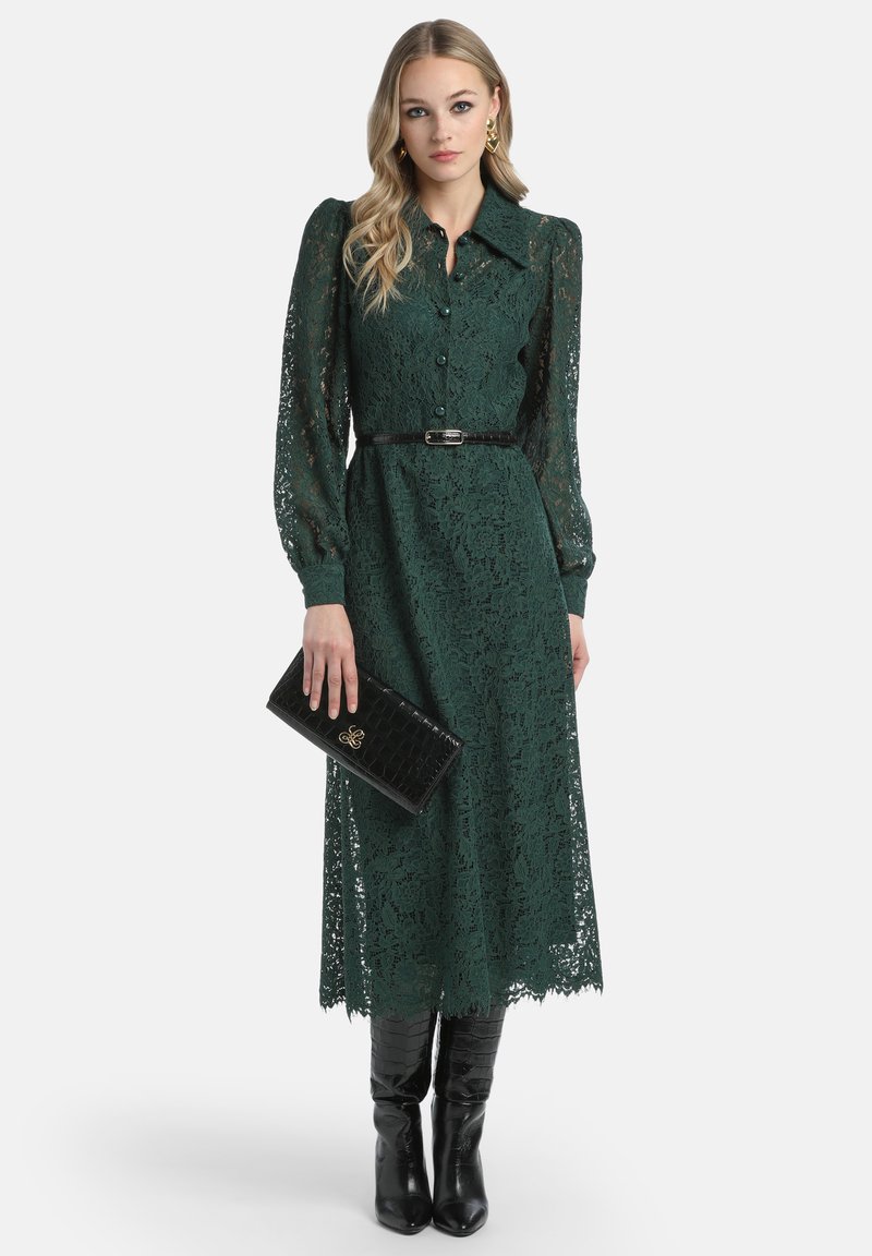 Grünes Spitzenkleid mit kragenförmigem Ausschnitt, langen Puffärmeln und tailliertem Gürtel. Kombiniert mit einer schwarzen Clutch aus Krokodilleder und knielangen Stiefeln.