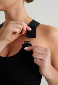 Černá sportovní podprsenka s nastavitelnými ramínky; modelova ruka odpojuje popruh na rameni. Hladký, elastický materiál.