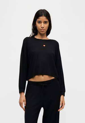 Jonge vrouw met lang donker haar draagt een zwarte cropped top met lange mouwen en zwarte broek, staand tegen een effen lichte achtergrond.