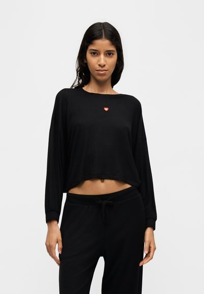 Млада жена с дълга тъмна коса носи черен къс топ с дълги ръкави и черен панталон, стои пред обикновен светъл фон.