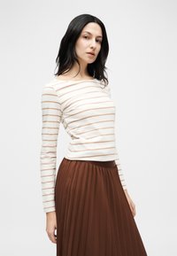 Γυναίκα με σκουρόχρωμα μαλλιά που φοράει κρεμ και καφέ ριγέ μακρυμάνικη μπλούζα και ψηλόμεση καφέ πλισέ φούστα σε λευκό φόντο.