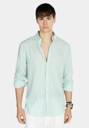 Joven con camisa de manga larga con rayas verticales en verde claro y blanco, con las mangas remangadas y pantalones blancos sobre un fondo liso.