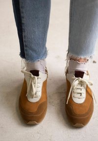 Sneakers in suede marrone con tomaia in pelle bianca, chiusura con lacci e dettagli testurizzati. Jeans in denim sfilacciato e calzini bianchi visibili.