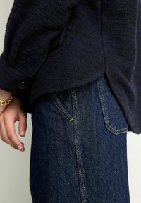 Navy textured fabric shirt with rolled sleeves, paired with dark denim jeans featuring gold stitching and angled front pockets.
