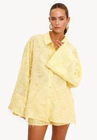 Chemise jaune texturée avec de grandes manches cloche et un col, dotée d'une fermeture boutonnée à l'avant et d'une surface duveteuse et inégale.
