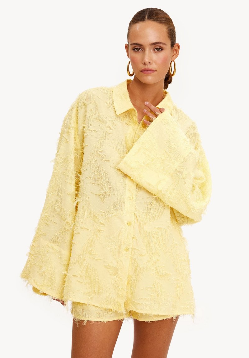 Chemise jaune texturée avec de grandes manches cloche et un col, dotée d'une fermeture boutonnée à l'avant et d'une surface duveteuse et inégale.