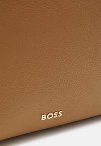 Hnědá kožená kabelka s texturovaným povrchem, na dolním pravém rohu s vyraženým zlatým logem "BOSS". Hladká a odolná.