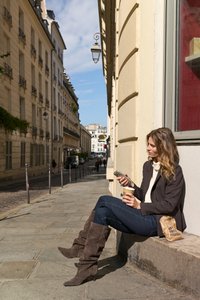 Γυναίκα κάθεται σε μια πέτρινη άκρη σε πεζοδρόμιο πόλης, κρατώντας ένα φλιτζάνι καφέ και κοιτάζει το τηλέφωνό της σε ηλιόλουστη μέρα.