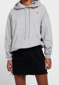Hoodie gris avec un cordon de serrage, arborant un petit logo rouge, associé à une jupe en denim noir avec un ourlet effiloché. Texture douce, design décontracté.