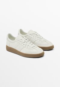 Baskets basses blanches avec semelles en caoutchouc beige texturées, à lacets, en matériau ressemblant au daim, présentées côte à côte sur un fond uni.