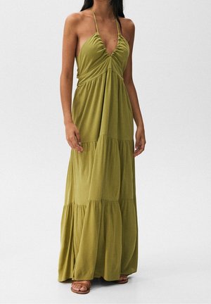 Vrouw draagt een mouwloze maxi-jurk in olijfgroen met gelaagde rokken en een halternek, samen met platte bruine sandalen, tegen een effen achtergrond.