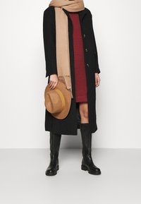 Manteau long noir avec boutons dorés, écharpe beige, robe bourgogne, chapeau marron et bottes noires jusqu'aux genoux, présentant des textures lisses et un design superposé.