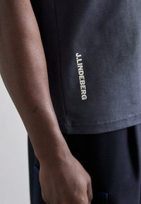 Siva majica kratkih rukava glatke teksture, sa bijelim logotipom "J.Lindeberg" na rubu, uparena s tamnim hlačama koje pokazuju plave akcentne boje.