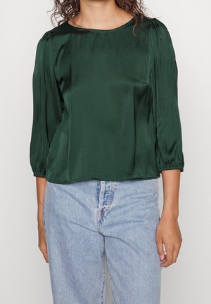 Kvinde iført en mørkegrøn satinbluse med trekvartærmer og lyseblå højtaljede jeans, stående foran en ensfarvet baggrund.