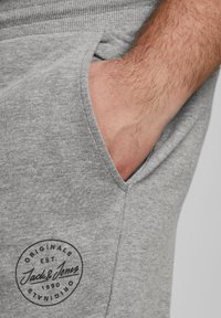 Pantalones de chándal grises con una tela texturizada, bolsillo lateral y un logo circular negro que presenta "Jack & Jones" y "Est. 1990."