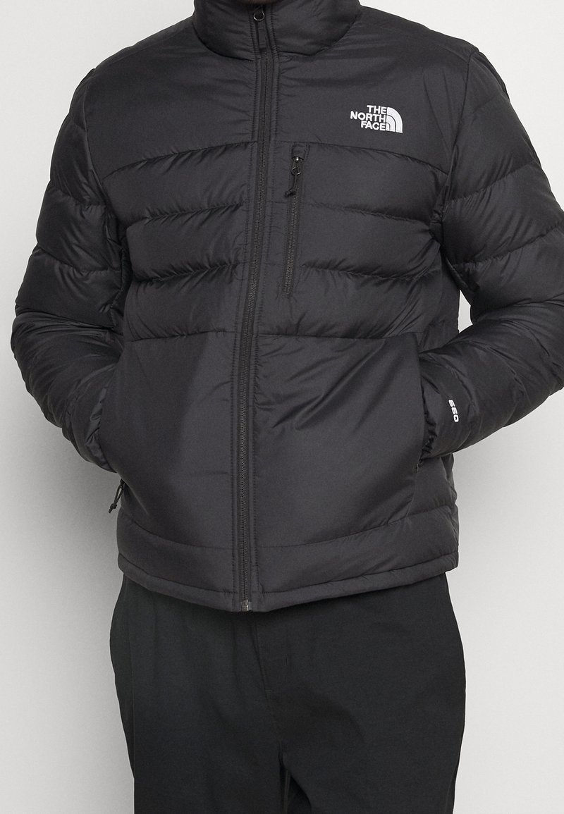 Man wearing a black The North Face quilted puffer jacket with zippered chest and side pockets, hands in pockets, standing against a plain background.