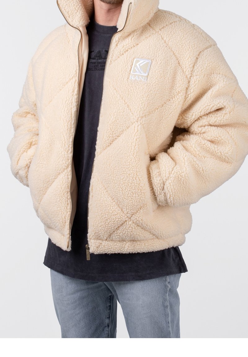 Beige schapenvachtjas met een ruitpatroon, ritssluiting en capuchon. Heeft een logo op de bovenste linkerkant van de borst. Gepaard met een zwart shirt.