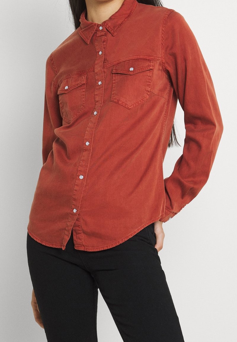 Camicia rossa a maniche lunghe con chiusura a bottoni, due taschini sul petto e orlo arrotondato. Realizzata in un tessuto morbido con una texture leggermente consumata.