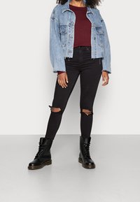 Lichtblauwe spijkerjas met lange mouwen en rafelige zoom. Daaronder een bordeauxrode top met lange mouwen, gecombineerd met zwarte skinny jeans met scheuren bij de knieën. Zwarte veterlaarzen met een dikke zool.