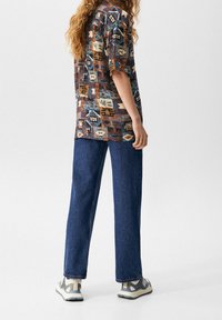 Chemise à manches courtes à motifs en marron, bleu et crème ; associée à un jean en denim bleu ample et des baskets grises avec des accents contrastants.